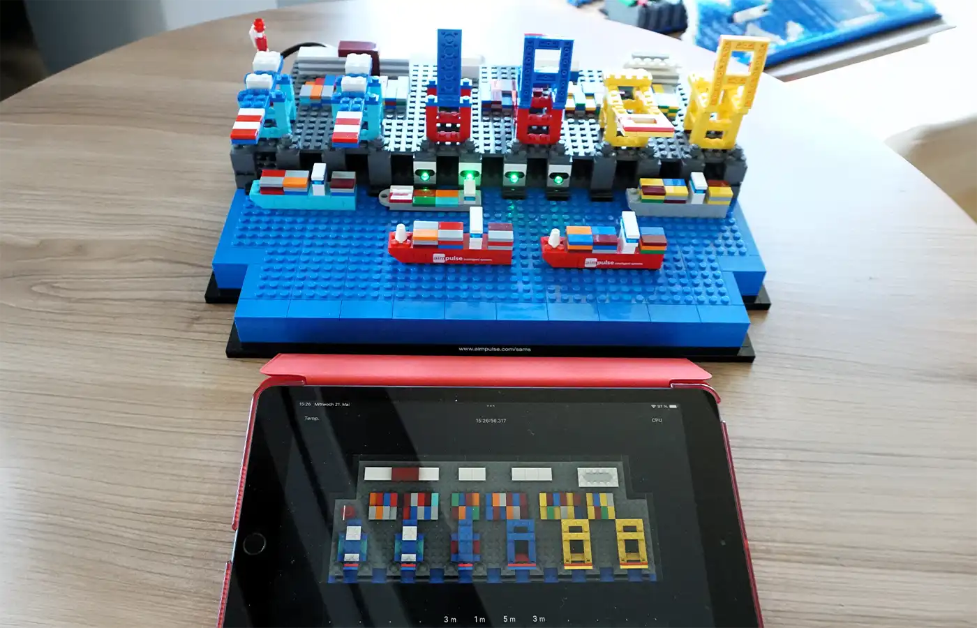 Ein aus Klemmbausteinen nachgebautes Hafenterminal mit Schiffen neben einem Tablet, das ein digitales Hafenlogbuch zur KI-gestützten Planung der Infrastrukturnutzung zeigt.