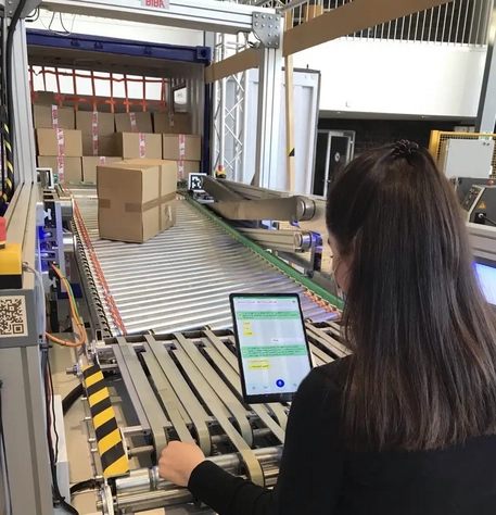 Mitarbeiterin bedient LogDynamics-Fördertechnik mit Tablet in einer automatisierten Lagerumgebung mit Paketen auf Rollenbahnen für COALA-KI-Projekte.