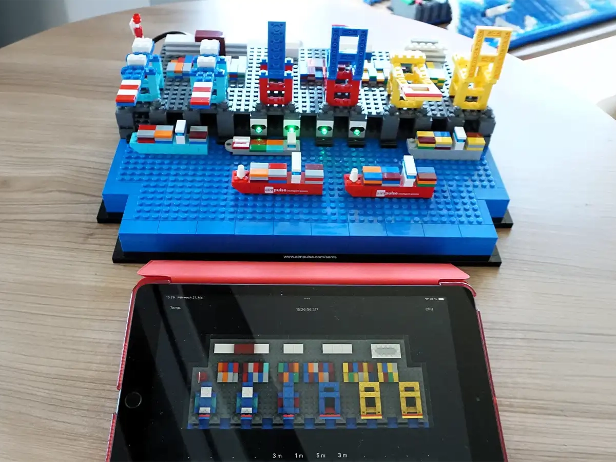Ein aus Klemmbausteinen nachgebautes Hafenterminal mit Schiffen neben einem Tablet, das ein digitales Hafenlogbuch zur KI-gestützten Planung der Infrastrukturnutzung zeigt.