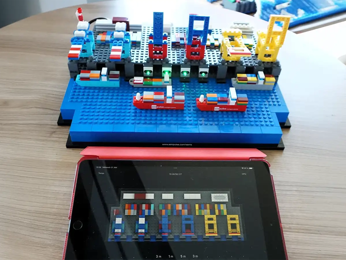 Ein aus Klemmbausteinen nachgebautes Hafenterminal mit Schiffen neben einem Tablet, das ein digitales Hafenlogbuch zur KI-gestützten Planung der Infrastrukturnutzung zeigt.